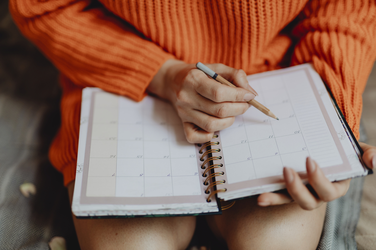 A person holds a planner notebook in their lap with one hand and a pencil with the other hand